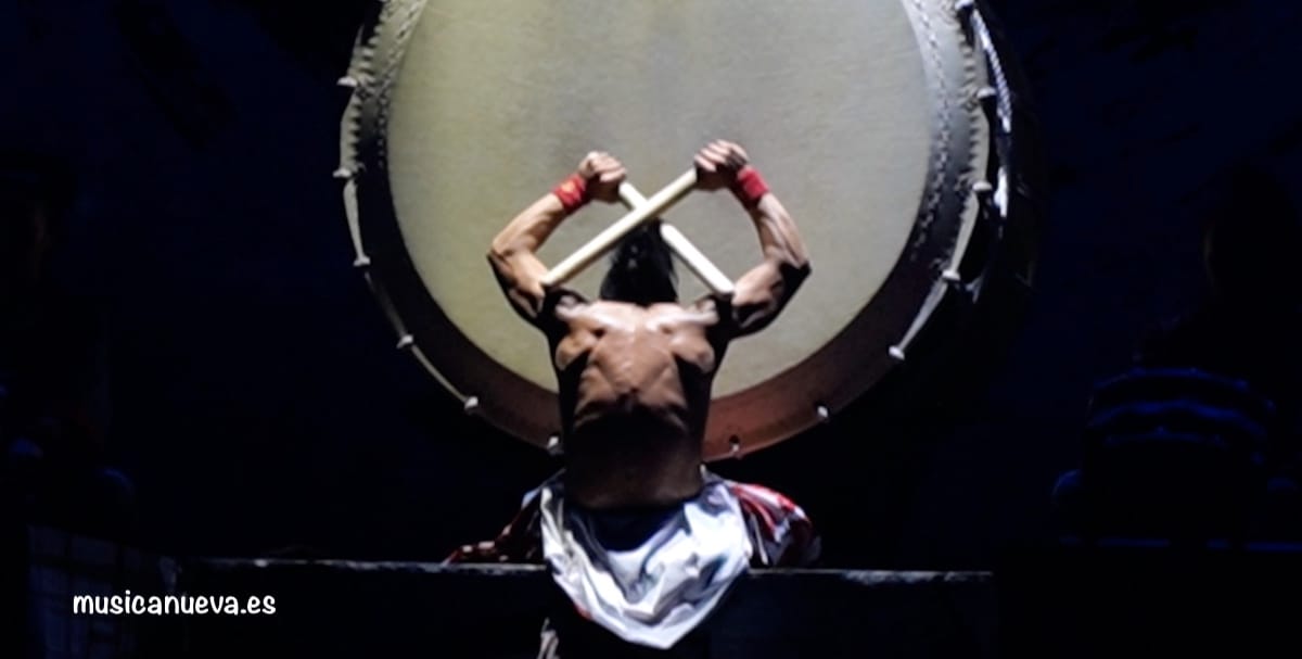 Un percusionista de taiko de espaldas muestra su musculatura mientras sostiene los palos sobre su cabeza frente a un tambor japonés gigante (O-daiko).