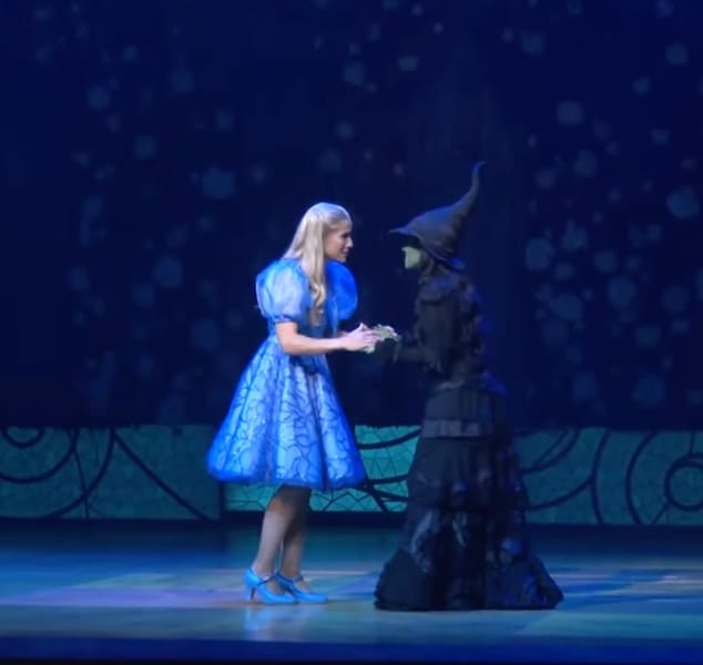 Glinda (vestida de azul claro) y Elphaba (de verde, vestida de negro y con sombrero de bruja) interactuando en el escenario durante una escena del musical WICKED.